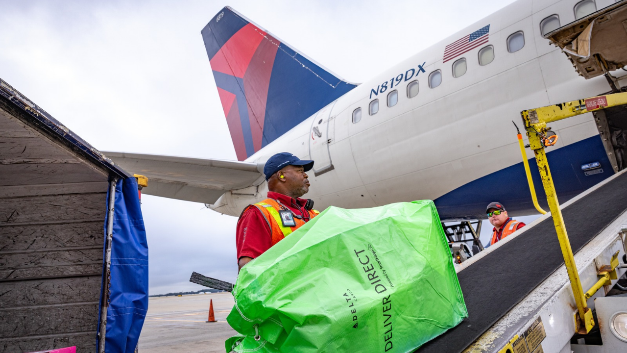 Delta ground crew member loading a green Deliver Direct cargo bag onto an aircraft at the airport